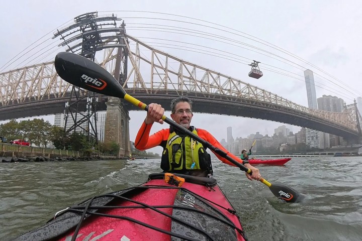 Two kayakers paddle on a river under a large suspension bridge with a cable car in the background.