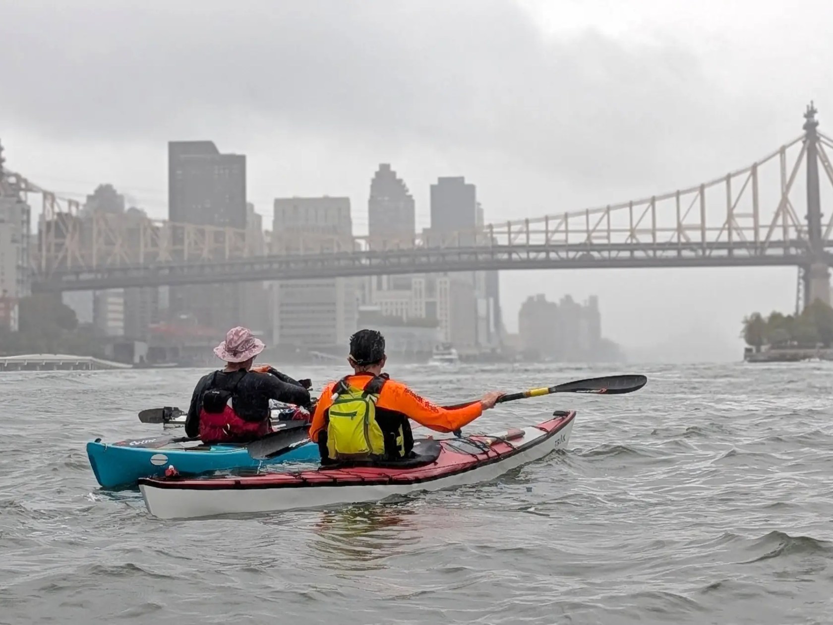Two kayakers paddle on a river with a cityscape and bridge in the background on a cloudy day.