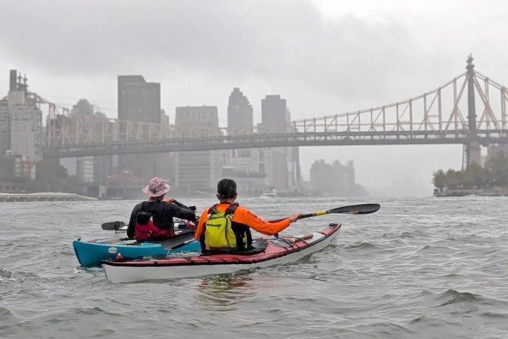 Two kayakers paddle on a river with a cityscape and bridge in the background on a cloudy day.