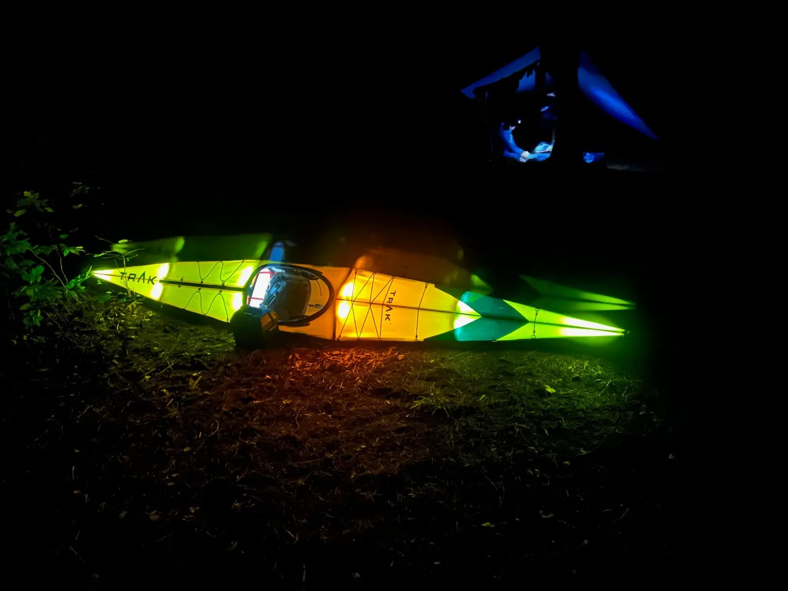 Illuminated kayak on ground at night with colorful lights, tent visible in the background.