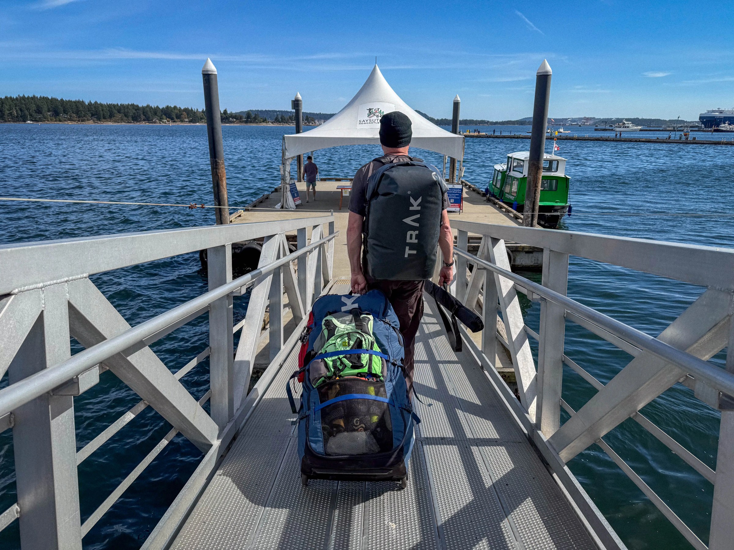Person with large backpack and gear on a dock walkway, near water and boats, under a clear sky.