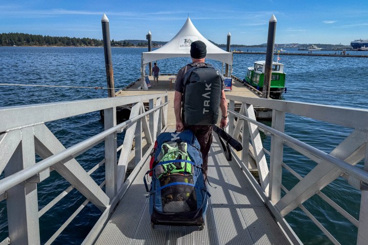 Person with large backpack and gear on a dock walkway, near water and boats, under a clear sky.
