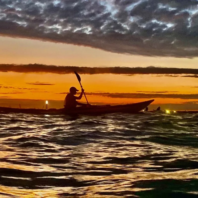 The sky ignites in deep orange as the sun sets over the Bayonne shipyards. A lone woman sea kayaker, silhouetted against the glow, uses a powerful high-angle stroke to navigate the choppy harbor water as her safety light flickers on.