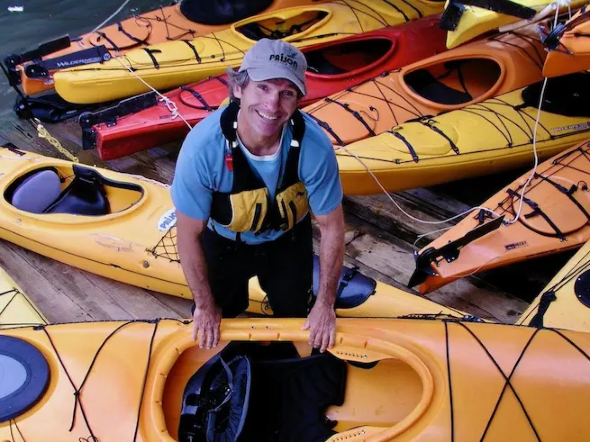 Person in a life jacket standing among numerous colorful kayaks on a wooden dock.