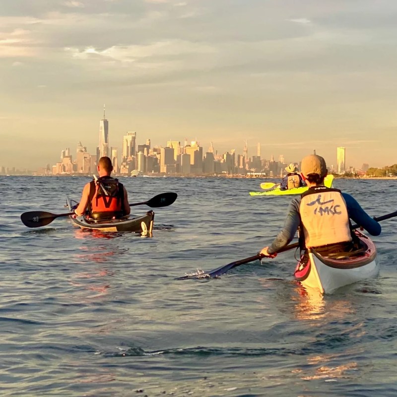 Two kayakers paddling toward the downtown Manhattan skyline in the early evening light.