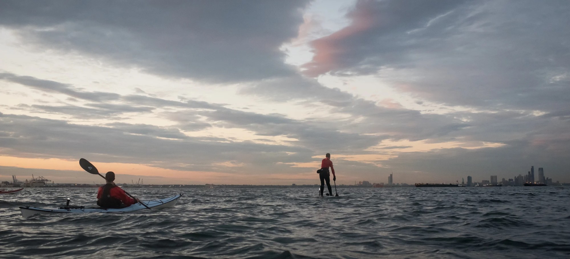 Kayaker and paddleboarder on water at sunset with city skyline in background.