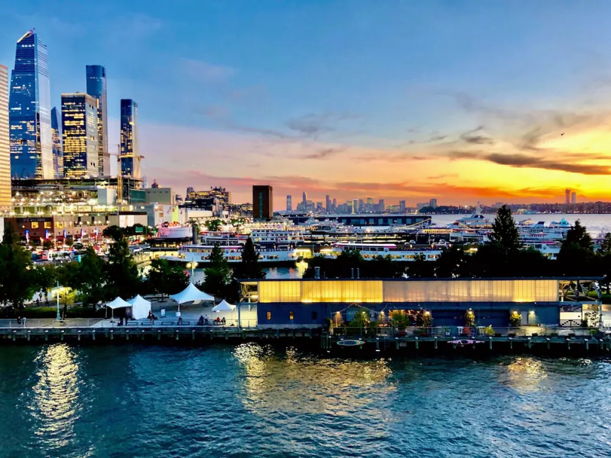 City skyline at sunset with skyscrapers, waterfront, and boats on the water.