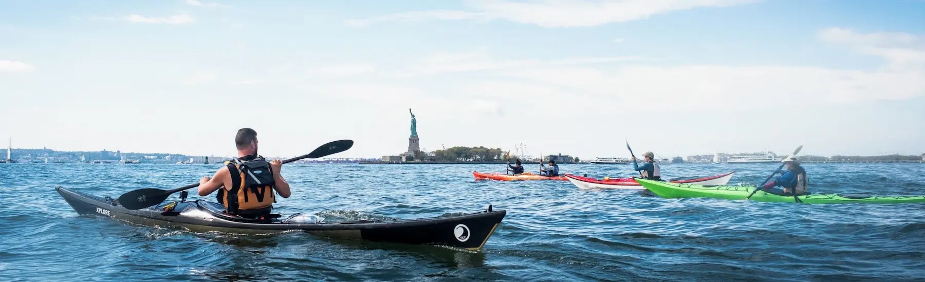 Four kayakers paddle through the open New York Harbor toward the Statue of Liberty, riding the ebb tidal current south on a bright, sunny day.