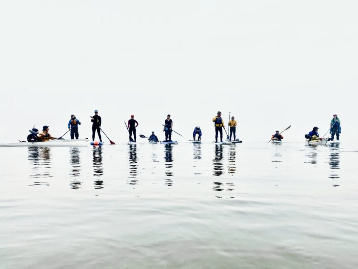 Group of people on paddleboards and kayaks on calm water against an overcast sky.