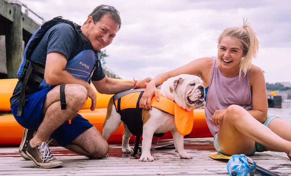 A candid, joyful moment at Manhattan Kayak: a smiling man and woman sit on the dock with a white English Bulldog wearing a bright orange dog lifejacket.