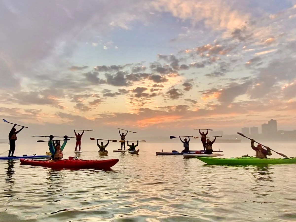 People in kayaks raising paddles against a vibrant sunset sky over calm water.