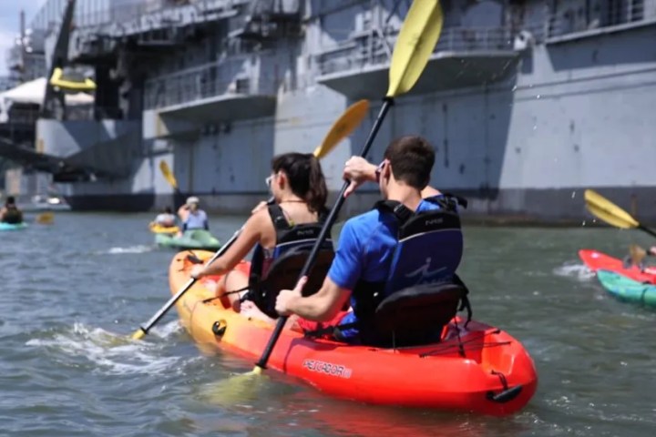 A couple doing a kayak rental in a bright orange tandem sit-on-top kayak along the massive grey hull of the Intrepid Museum.