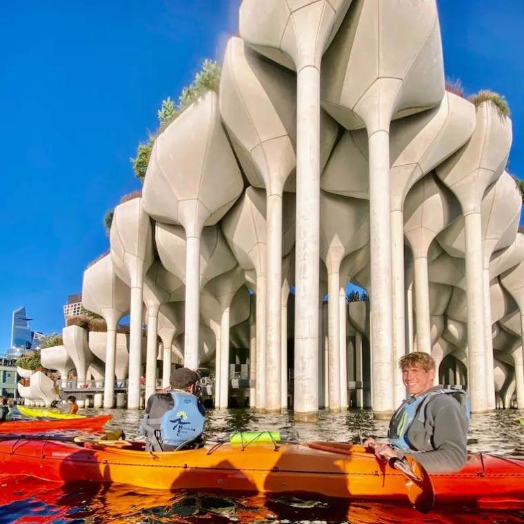 People sea kayaking on the Hudson River alongside Little Island with large, sculptural columns under a clear blue sky.