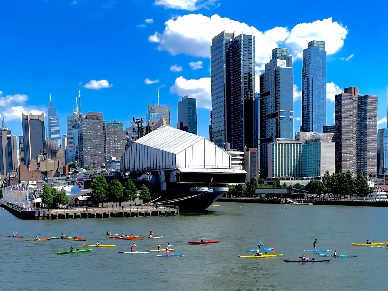 A vibrant community of 23 Manhattan Kayak Co sea kayakers and paddleboarders explore the Hudson River outside of the Pier 84 Boathouse in Hudson River Park, with the USS Intrepid and Hudson Yards skyline in the background.
