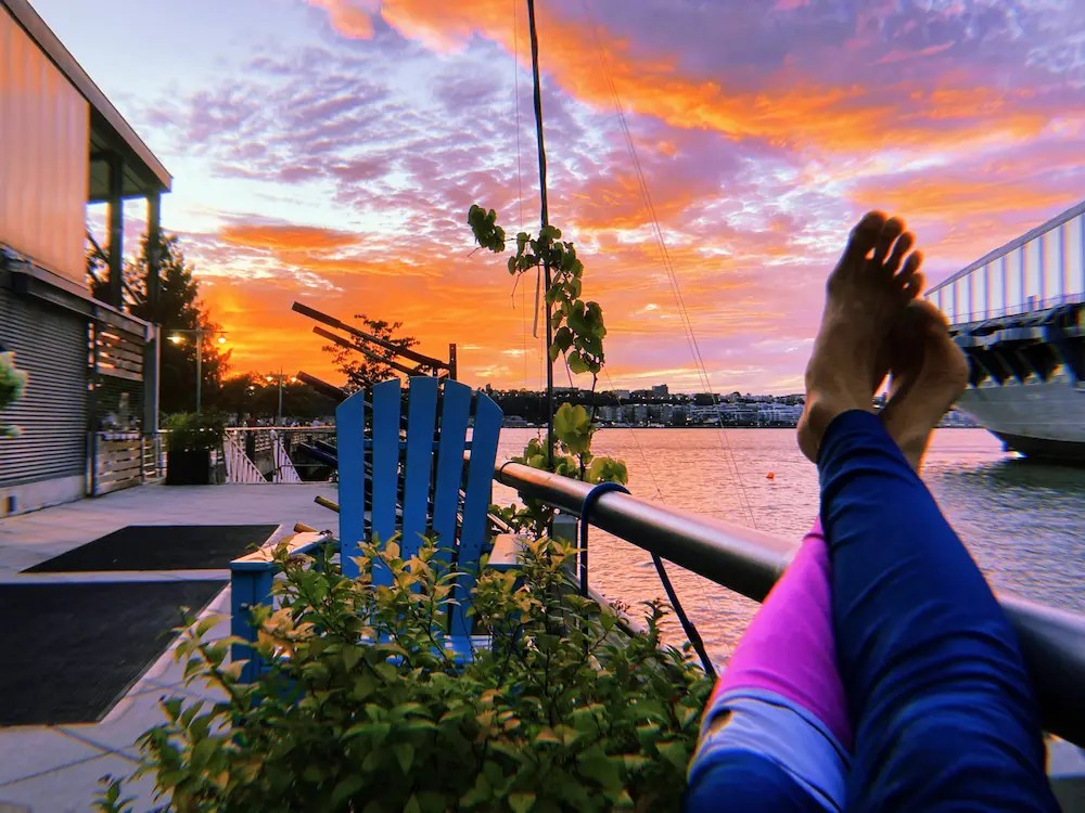 A Manhattan Kayak Co's Adventure Club member relaxes on the waterfront patio of the Pier 84 Boathouse. Her feet are crossed at the ankles and propped up on table as vibrant orange and purple sunset clouds roll over the sky.