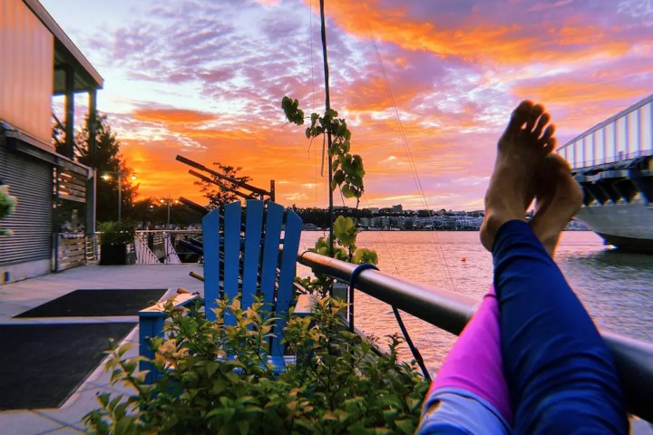 A POV of feet, crossed at the ankles, propped up on table at the Pier 84 Boathouse waterfront patio, as vibrant orange and purple clouds dominate the sky.