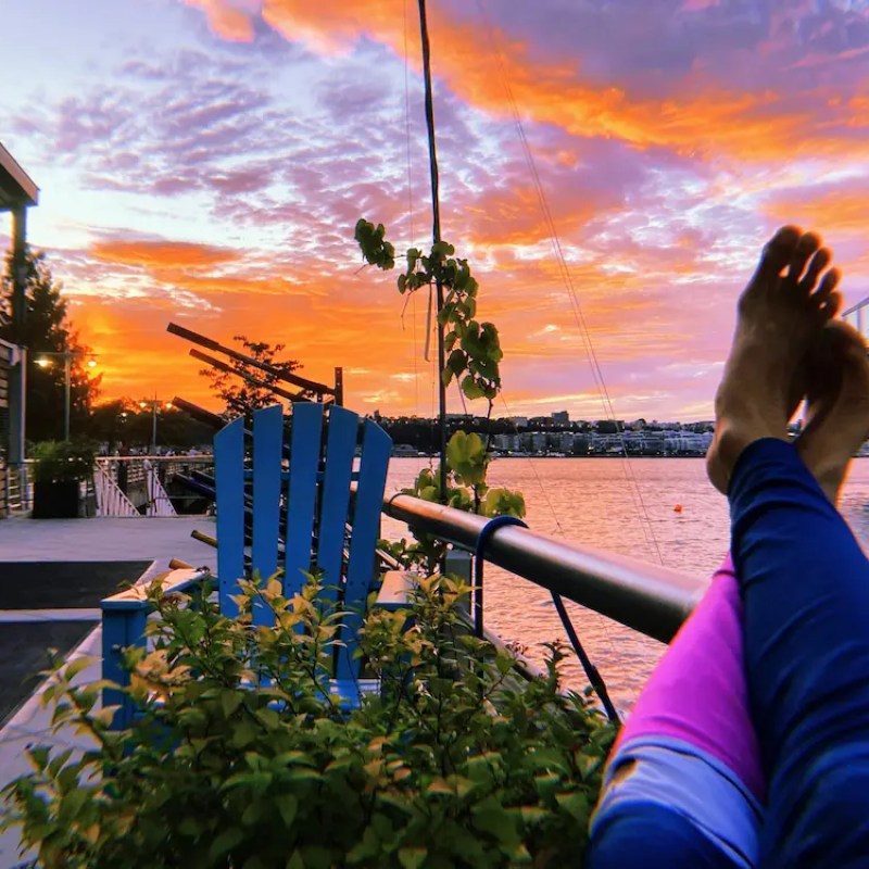 A Manhattan Kayak Co's Adventure Club member relaxes on the waterfront patio of the Pier 84 Boathouse. Her feet are crossed at the ankles and propped up on table as vibrant orange and purple sunset clouds roll over the sky.