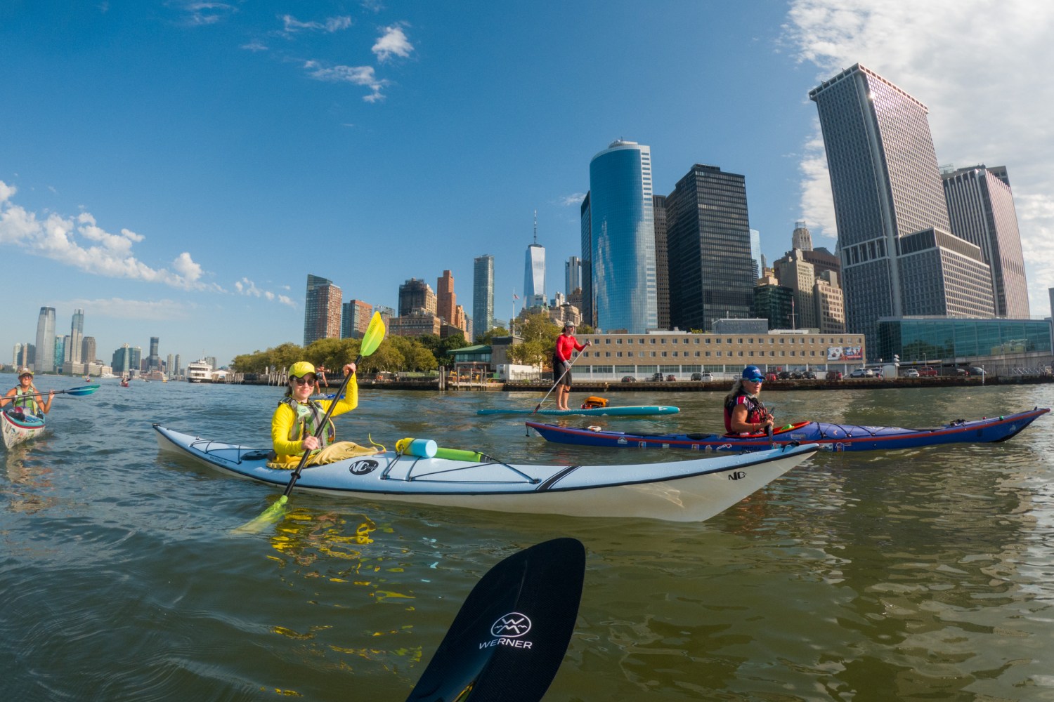 People kayaking on a river with a city skyline in the background under a clear blue sky.