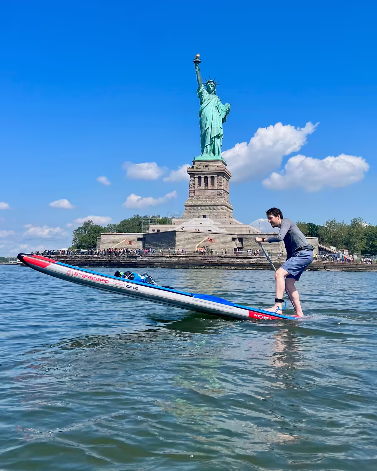 A man raising the nose of a Starboard SUP entirely out of the water while maintaining balance in front of the Statue of Liberty. This step-back pivot turn is an advanced maneuver requiring precision, core strength, and technical mastery. Most people see the Statue of Liberty from a crowded ferry; paddling there is the ultimate secret NYC experience. There are crowds of tourists on the pedestal of the statue in the background. The contrast between the masses and the solo athlete on the water is a powerful status narrative for a high-achieving professional. The deep Lady Liberty green, the crisp blue sky, and the pops of red and blue on the board create a vibrant, classic American color story.