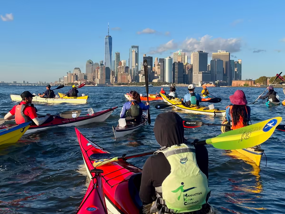 A group of 15+ high-end touring kayaks in the middle of the NY Bay looking north toward the Lower Manhattan skyline and One World Trade Center. There is a high-energy athletic vibe where paddlers push their limits alongside peers who share that drive. The primary colors of the boats (yellow, red, blue) pop against the deep blue water and the crisp, clear sky. The way the group fans out toward the city creates a powerful sense of forward momentum and collective purpose.