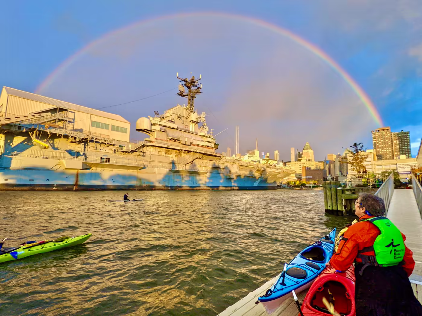 A vibrant rainbow perfectly frames the Intrepid Museum. A paddler, who has just come back from a river trip, is looking at the rainbow with mindfulness and presence. This awe-inspiring moment is the ultimate luxury in NYC.