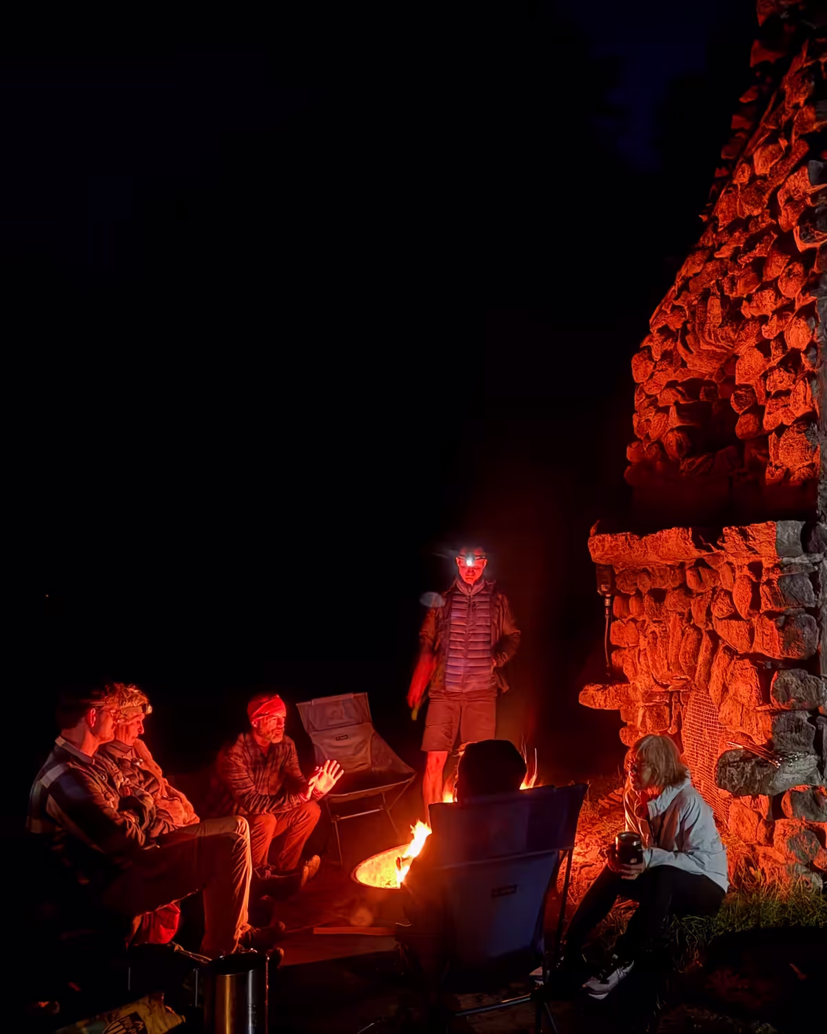During an away trip, a group of members gathers around a campfire by the towering ruins of a massive stone fireplace. This is an immersive backcountry camping trip, a world away from the city's concrete. The scale of the stone chimney provides an almost gothic backdrop. The warm, orange glow of the fire illuminates the paddlers’ faces and the weathered stonework against the pitch-black night. The group wears headlamps and technical down jackets. After a long day of paddling to the campsite, this is their recovery phase. This builds the kind of brotherhood and community that is difficult to find in a city full of distractions.