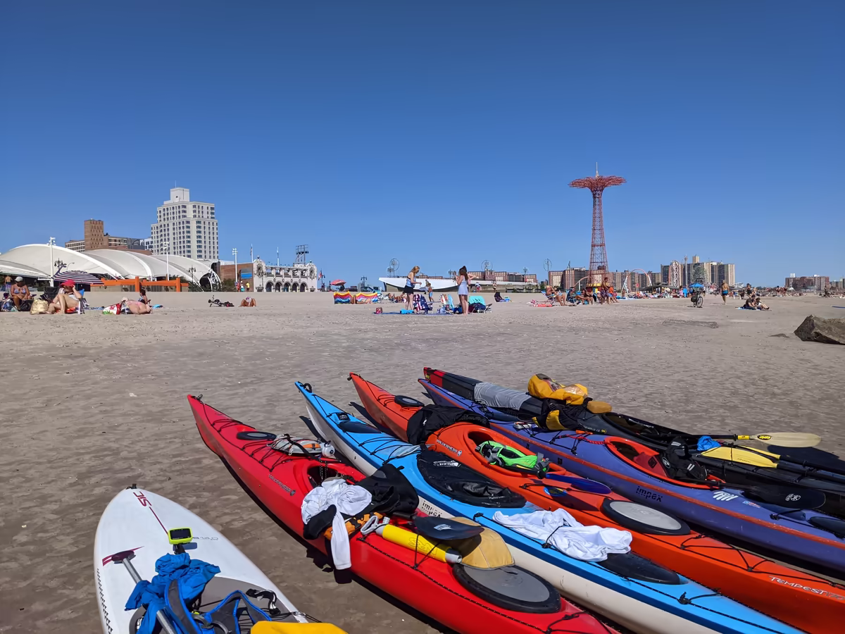 A fleet of Manhattan Kayak Co's performance sea kayaks (including P&H Cetus, Wilderness Systems Tempest, and Impex models) and a SIC Maui racing SUP parked on Coney Island Beach, with dry bags and paddles ready for the 17-mile return leg back to Manhattan. The iconic Parachute Jump and Ford Amphitheater are in the background. While millions of people visit Coney Island by subway, this tribe arrives via the Verrazzano Narrows and the Atlantic Ocean.