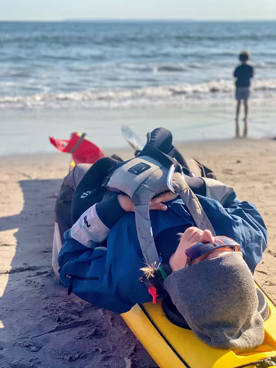 A woman wearing a beanie, sunglasses, lifejacket, and kayaking gear is lying on top of her sea kayak on Coney Island Beach. She is in deep relaxation.