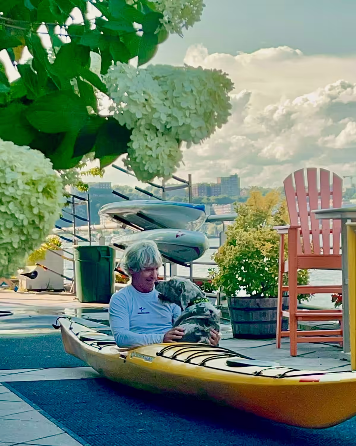 A candid moment of connection between Eric Stiller, Founder of Manhattan Kayak Co, sitting in a yellow touring kayak with his dog and paddle companion Charlie, on the pier at Manhattan Kayak Co. (Pier 84). Eric dared to create a world where he can enjoy his dog, his sport, his city, and his friends altogether. All who enter are welcome to live that lifestyle too. The lush white hydrangeas in the foreground create a soft, natural frame. The lighting is warm, diffused, and serene.
