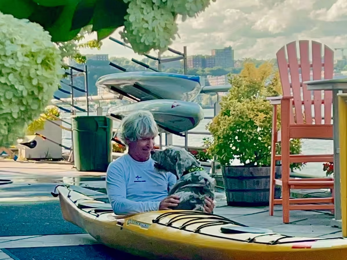 A candid moment of connection between Eric Stiller, Founder of Manhattan Kayak Co, sitting in a yellow touring kayak with his dog and paddle companion Charlie, on the pier at Manhattan Kayak Co. (Pier 84). Eric dared to create a world where he can enjoy his dog, his sport, his city, and his friends altogether. All who enter are welcome to live that lifestyle too. The lush white hydrangeas in the foreground create a soft, natural frame. The lighting is warm, diffused, and serene.