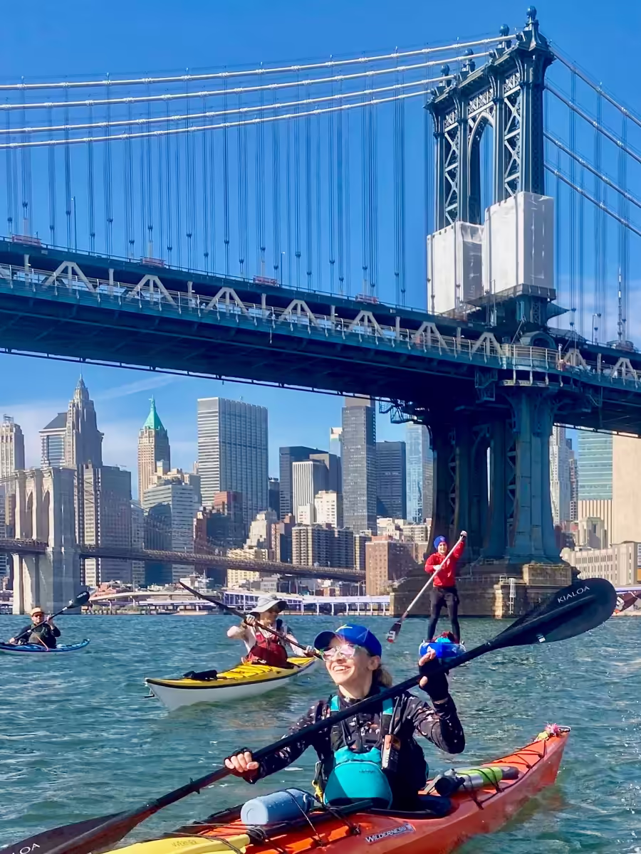 A group of Manhattan Kayak Co paddlers on the East River during the 30-mile expert-level Manhattan Circumnavigation trip. They just passed under the Manhattan Bridge. The Brooklyn Bridge and Wall Street skyline are visible in the background.