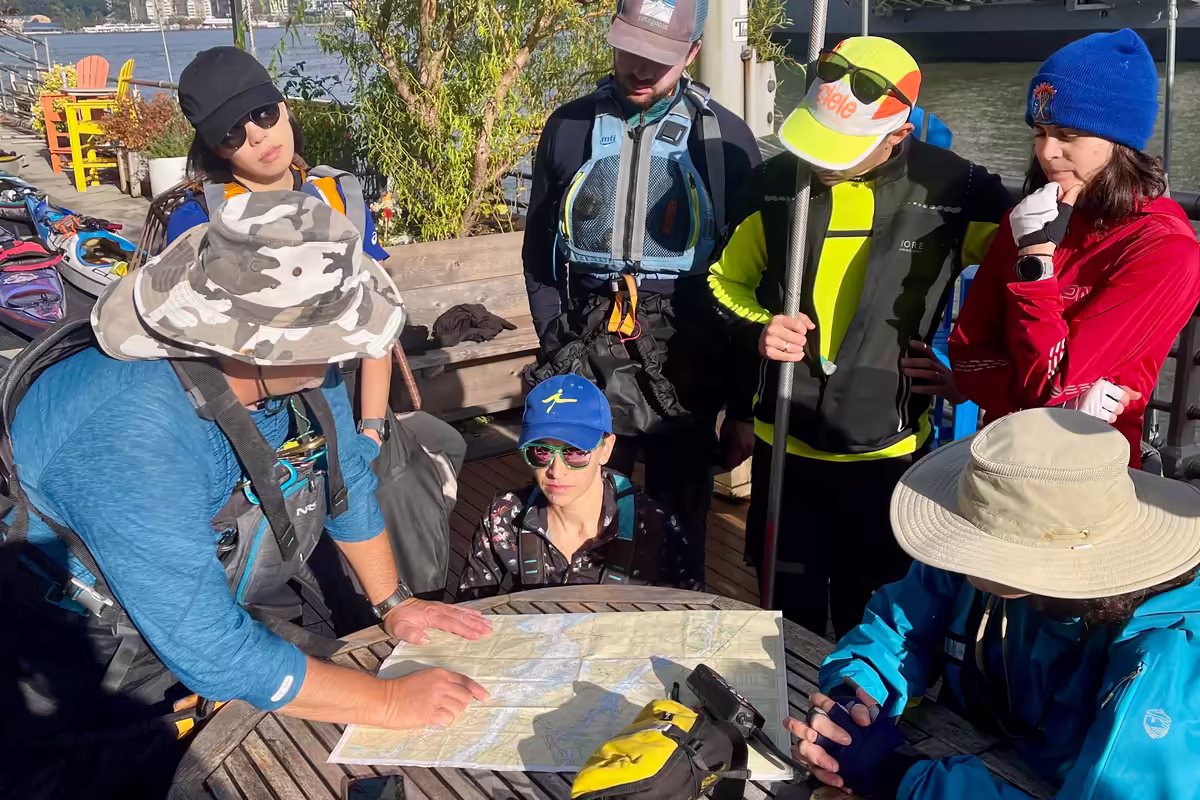 You can feel the concentration and the pre-trip adrenaline surrounding this group of kayakers, who huddle around a weathered wooden table on the outdoor deck of the Pier 84 Boathouse in the morning sunlight. The guide is pointing to a physical nautical chart of the New York Harbor. Everyone is kitted out in high-end technical apparel—life jackets, brimmed hats, and moisture-wicking layers. They are about to navigate the waters of a global megacity. It’s a frontier experience hidden in plain sight.