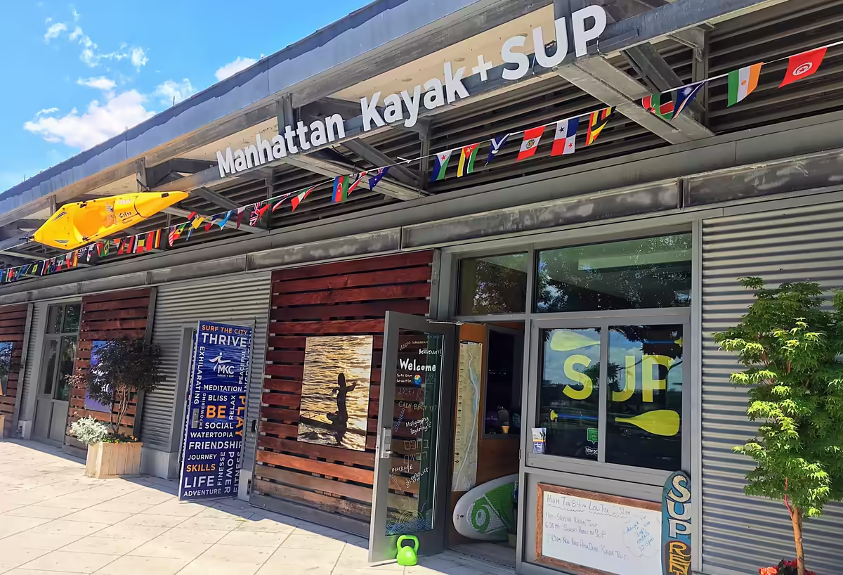 This is the front entrance to the Pier 84 Boathouse in Hudson River Park, where Manhattan Kayak Co runs a professional public paddling center offering low-cost rentals, classes, and guided trips for all skill levels. The entrance is adorned with a colorful string of flags and a yellow kayak above blue double doors and wooden sliding doors. The welcoming and organized entrance leads to a high-quality setting for groups and visitors.
