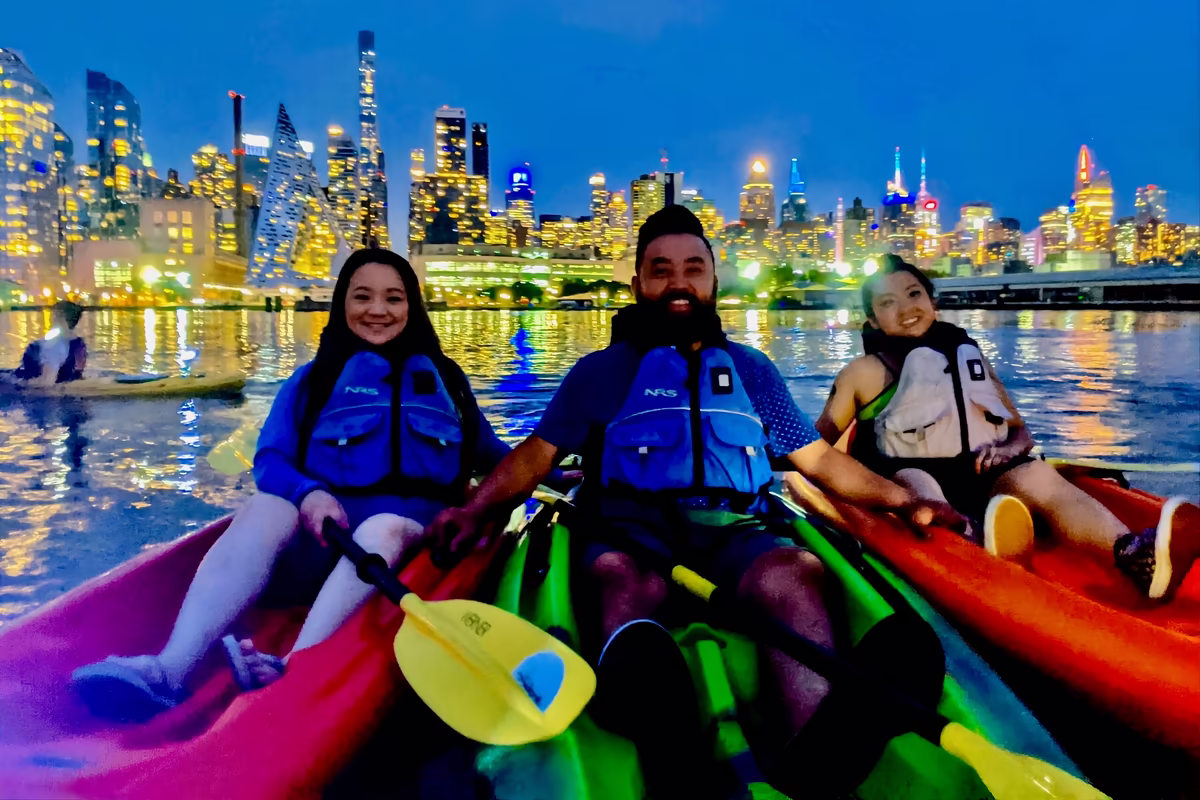 Three friends in glowing LED-lit kayaks smile on the Hudson River, with the illuminated Midtown Manhattan skyline and Hudson Yards glowing behind them during a night kayak tour.