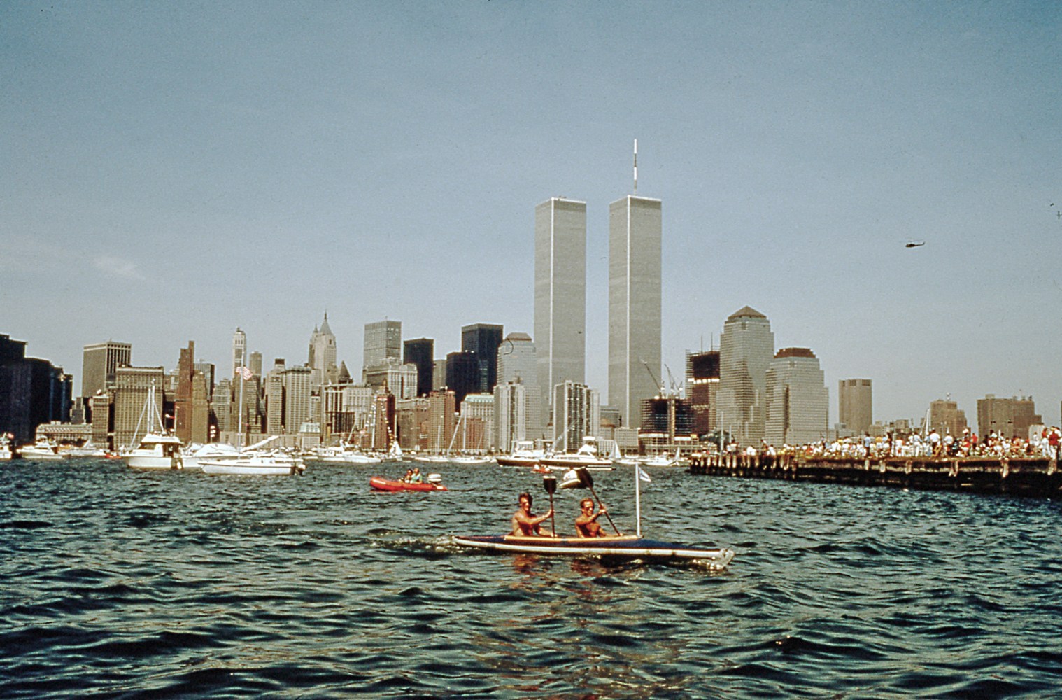 Two people kayaking with New York City skyline, including Twin Towers, in background.