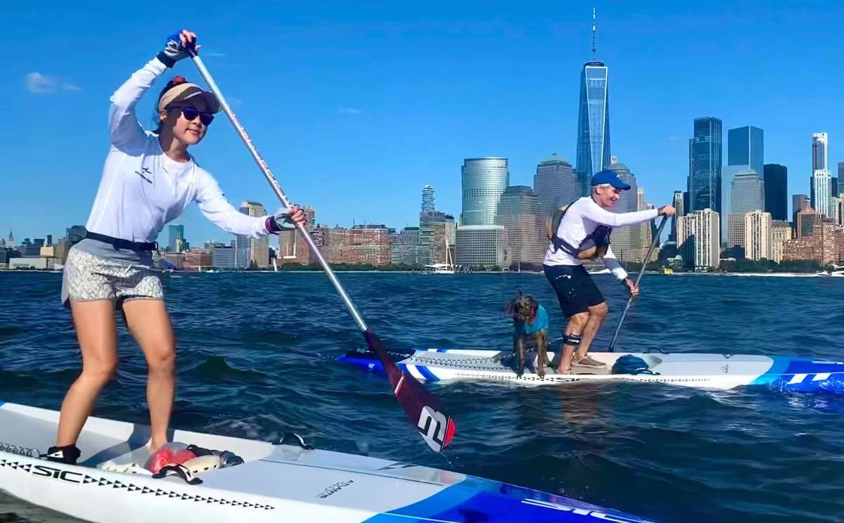 This is the work-hard, play-hard spirit alive in a female athlete and a male athlete with a dog, both riding SIC Maui’s Atlantis high-performance racing SUPs, with the Lower Manhattan skyline, One World Trade, and Goldman Sachs headquarters at 200 West Street in the background, and a crisp blue sky beyond. Both paddlers demonstrate excellent technical form.