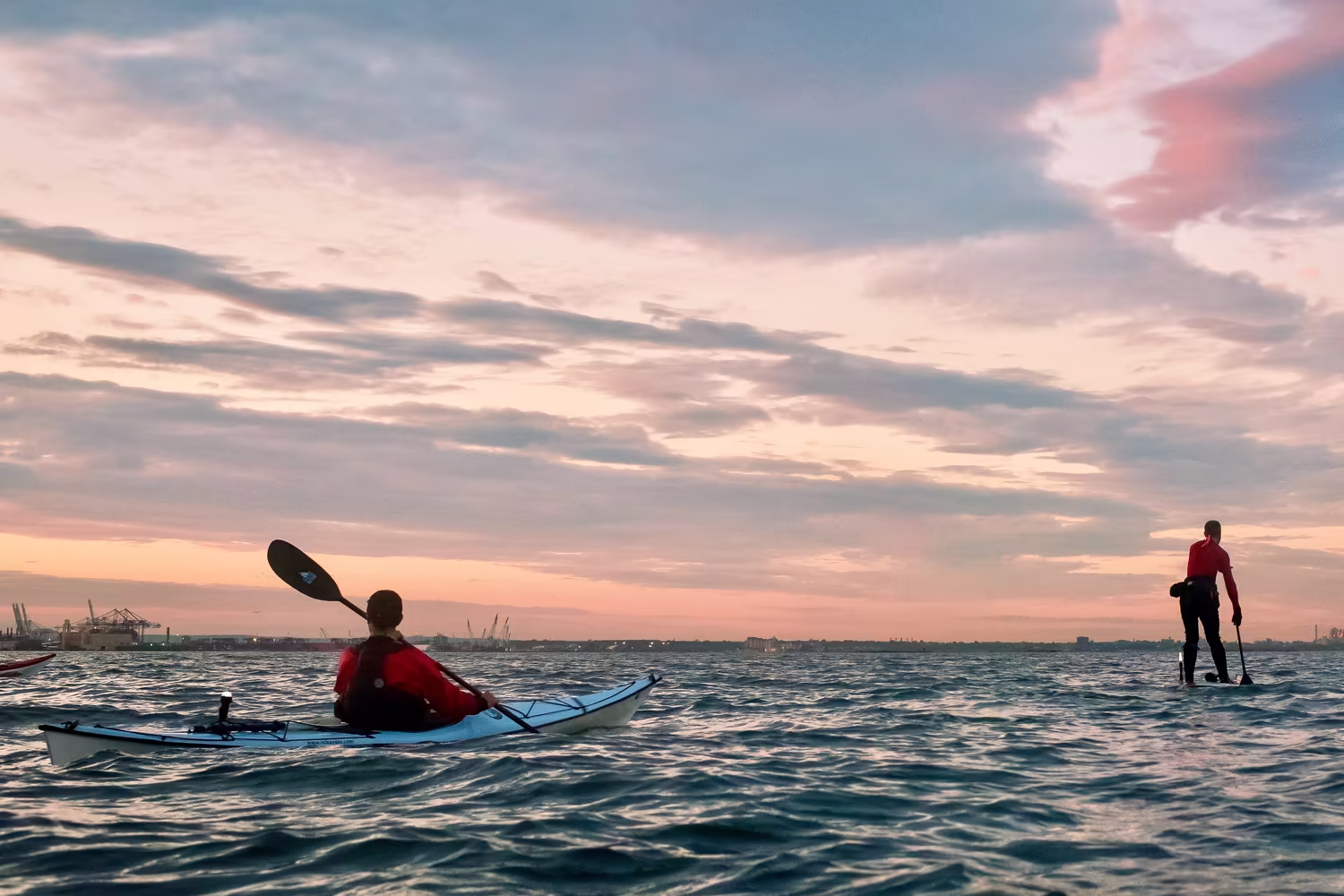 Under a soft, expansive arch of pale pink clouds, two tiny figures—a kayaker and a paddleboarder—look like specks against the vastness of the Upper Bay. They pull toward a distant Manhattan skyline, while the Statue of Liberty and Jersey City appear as mere flickers on a low, hazy horizon. Photo by Regina Nicolardi.