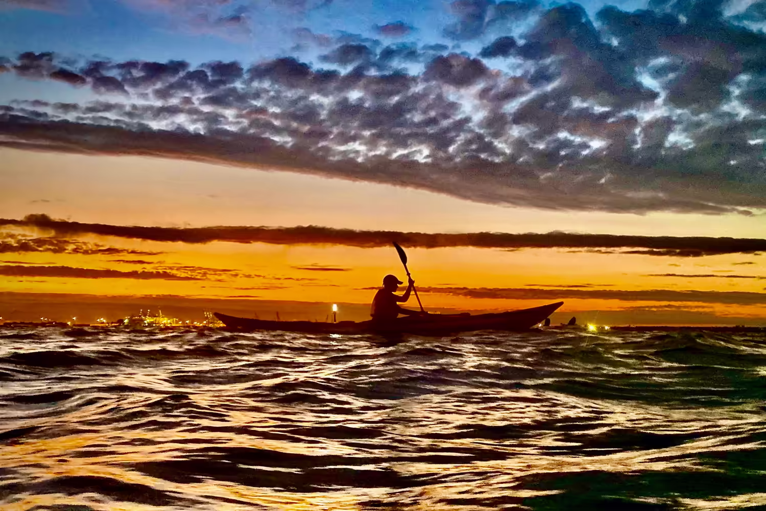 A lone kayaker is carved in black silhouette against a fiery, copper-orange sky as the sun dips behind the industrial cranes of Bayonne. She executes a sharp, determined stroke to cut through the dark, choppy harbor water, while rippled mackerel clouds catch the last of the intense light overhead. Photo by Regina Nicolardi.