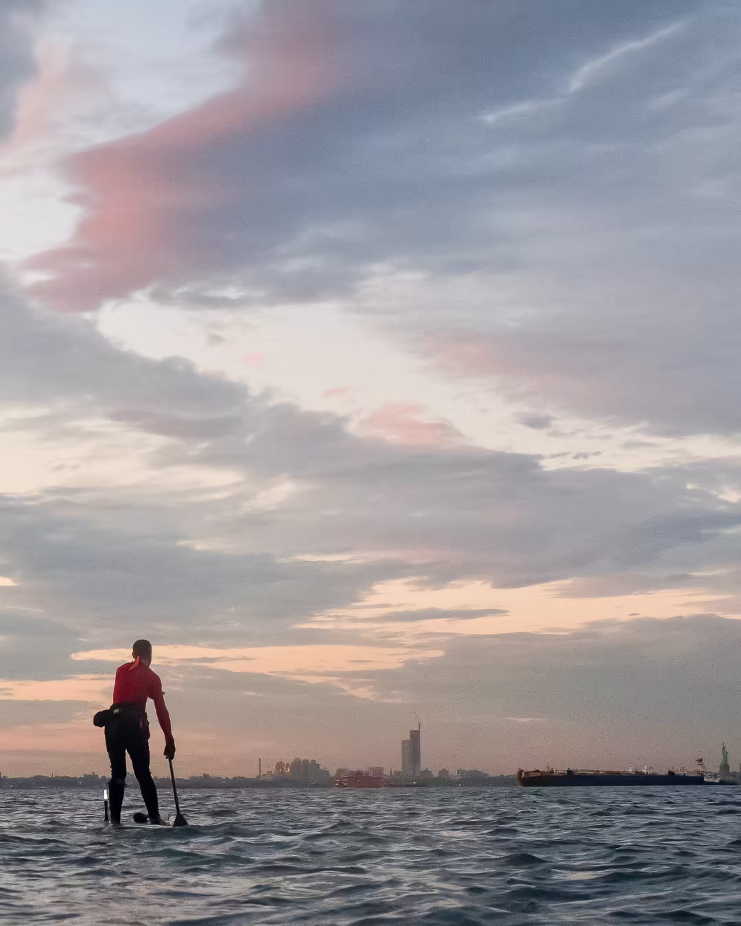 A paddleboarder heading home, traversing the vast Upper Bay toward Manhattan. The Statue of Liberty and Jersey City are tiny flickers on the horizon under pale pink clouds.