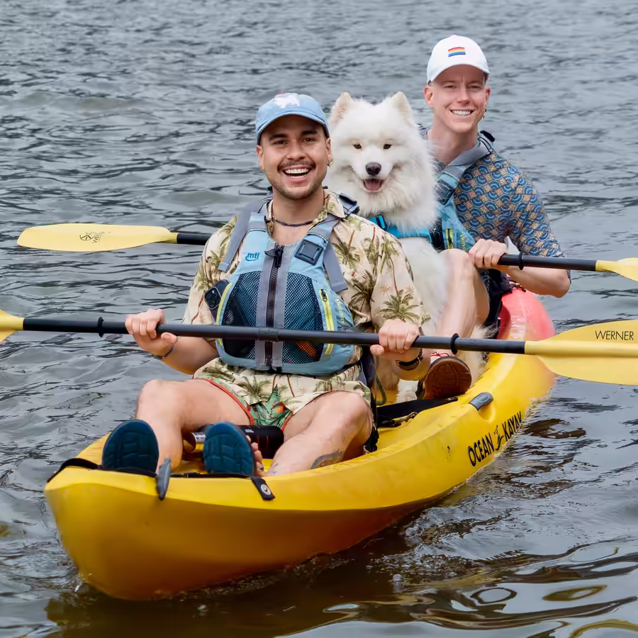 Two men paddling a tandem kayak with a large Samoyed dog sitting in the middle. Stable dog-friendly kayak rentals at Manhattan Kayak, Pier 84, showing how our wide boats easily accommodate large dogs and multiple paddlers.