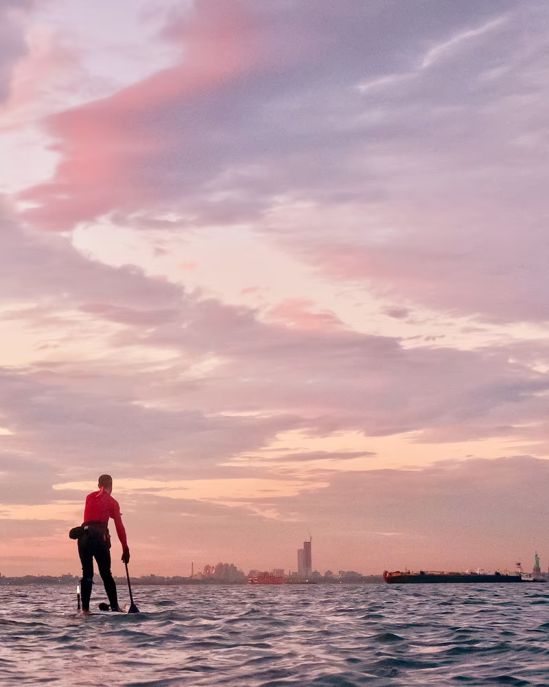 Under a soft, expansive arch of pale pink clouds, one tiny figure—a stand-up paddleboarder—looks like a speck against the vastness of the Upper Bay. He pulls toward a distant Manhattan skyline, while the Statue of Liberty and Jersey City appear as mere flickers on a low, hazy horizon. Photo by Regina Nicolardi.