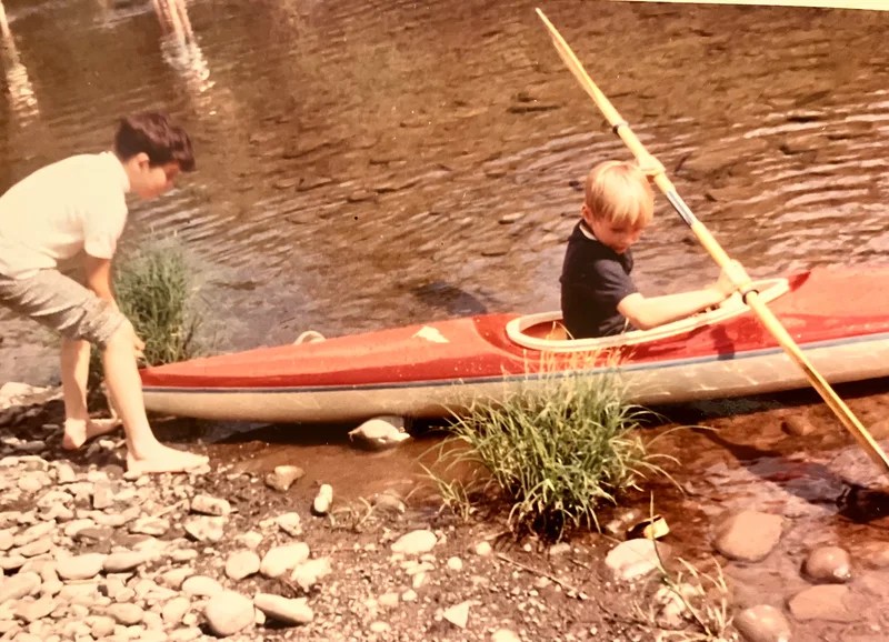 Two children with a red kayak on a rocky riverbank.