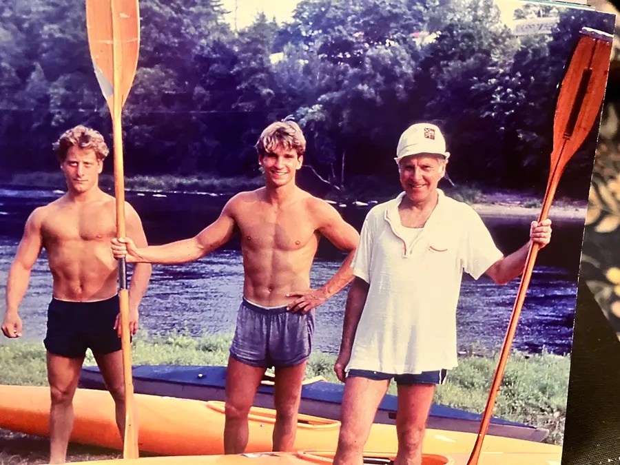 Three men with oars by a river, smiling, wearing shorts and casual tops.