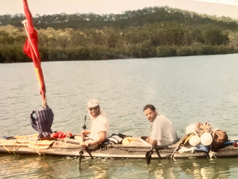 Two people kayaking on a lake with forested hills in the background.