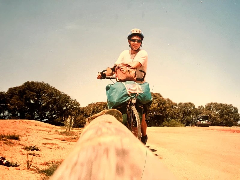 Person biking on a sunny day, carrying bags, trees and car in background.