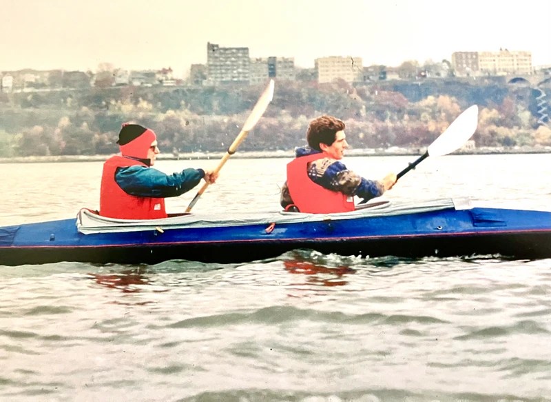 Two people kayaking on a river with city buildings in the background.