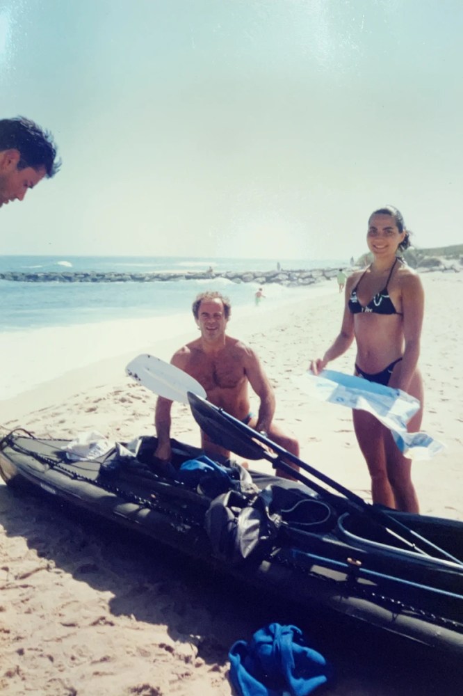 Three people with a kayak on a sunny beach, with one man kneeling and a woman holding a towel.