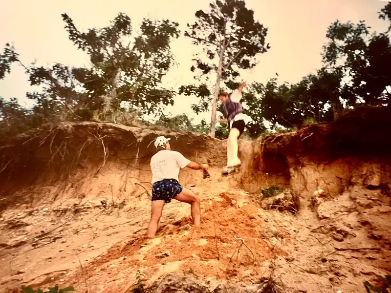 Two people climbing a steep sandy hill with trees above.