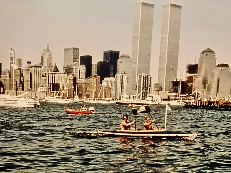 Two people canoeing on water with cityscape and Twin Towers in background.
