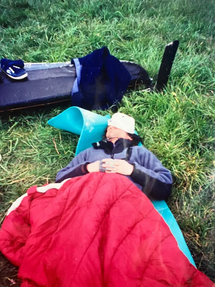 Person sleeping on a mat outdoors with a red sleeping bag and blue jacket.
